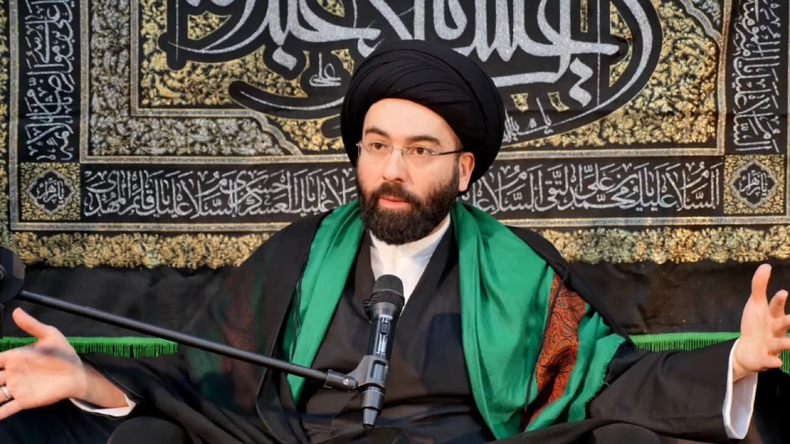 Sayed Mahdi Modarresi teaching before a calligraphic banner.